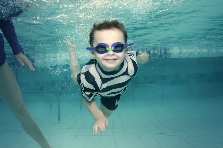 A young child swimming underwater towards the camera, smiling behind goggles.